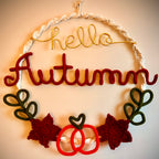 A handmade autumn-themed wall hanging with a macrame hoop, a gold wire script reading 'hello', a large red felt word 'Autumn', dark green crocheted leaves, and red and white decorative elements at the bottom, shown against a plain beige wall.