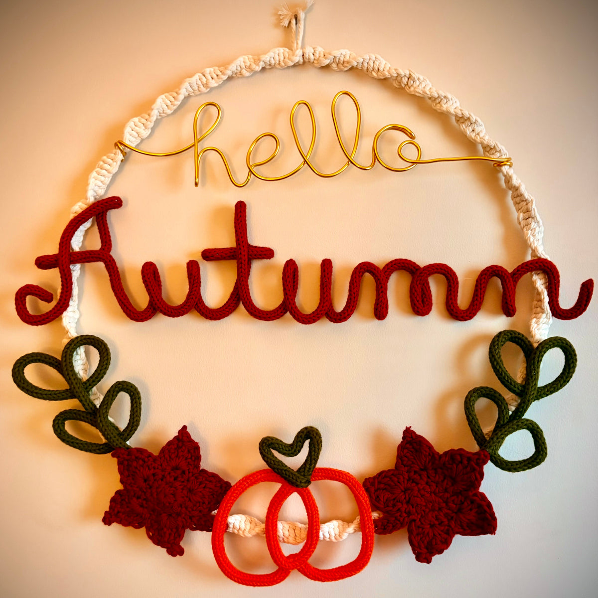 A handmade autumn-themed wall hanging with a macrame hoop, a gold wire script reading 'hello', a large red felt word 'Autumn', dark green crocheted leaves, and red and white decorative elements at the bottom, shown against a plain beige wall.